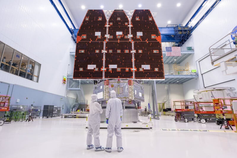 Two people in white cleanroom suits stand in front of a large spacecraft with folded solar panels inside a bright, spacious laboratory filled with equipment, ladders, and an American flag on the wall.