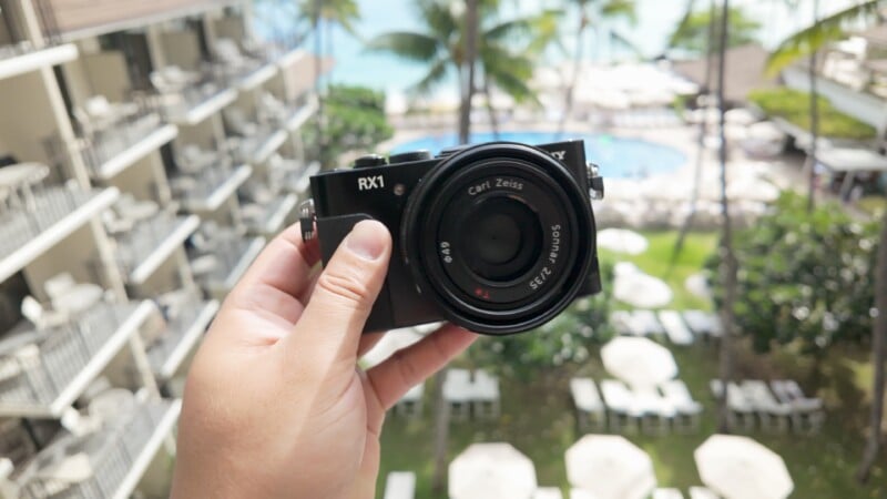 A hand holds a black RX1 camera with a Carl Zeiss lens, positioned above a hotel balcony overlooking a pool, palm trees, and lounge chairs in a sunny outdoor setting.