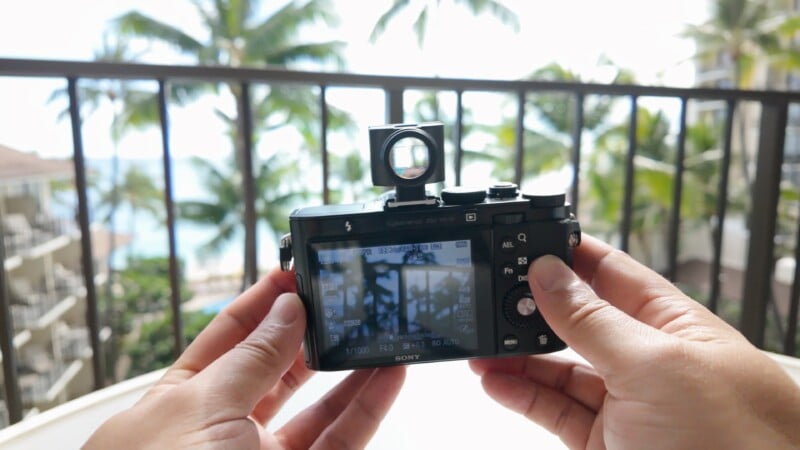 A person holds a digital camera with a flip-up screen showing a view of palm trees, balcony railings, and a bright, tropical outdoor scene in the background.