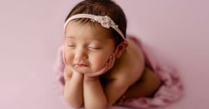 A baby with closed eyes rests their chin on their hands, lying on a soft pink blanket. The baby wears a light pink headband with a small flower, and the background is a pale pink shade.