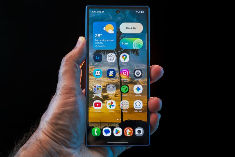 A hand holds a smartphone displaying a home screen with app icons, a weather widget showing 28°C and "Rain ending around 6:45 AM," and a beach sunset wallpaper with palm trees.