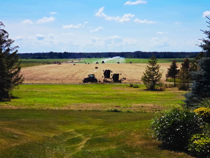 A tractor works in a large, harvested field under a blue sky with scattered clouds. Sprinklers water the field in the distance, and trees border the area, with green grass and bushes in the foreground.