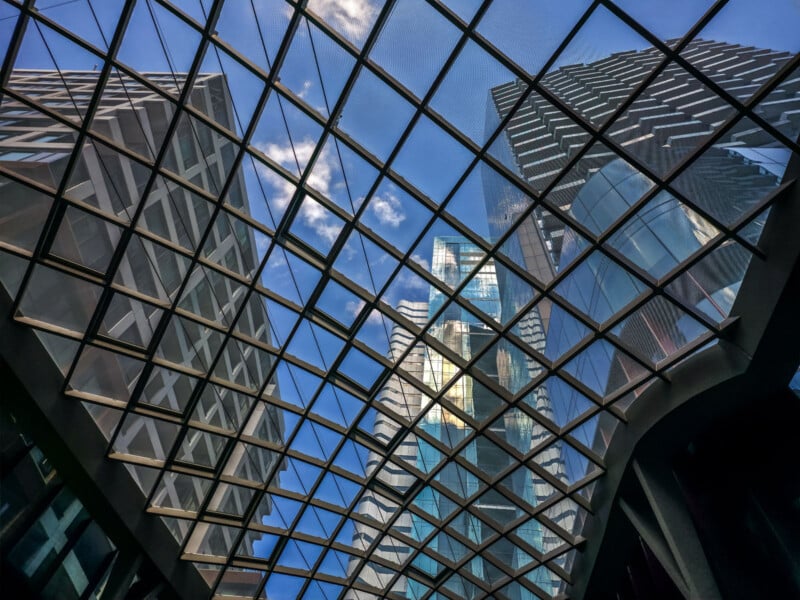 View of modern skyscrapers through a geometric glass roof grid, looking up at blue sky and clouds with reflections on the glass and tall buildings surrounding the structure.
