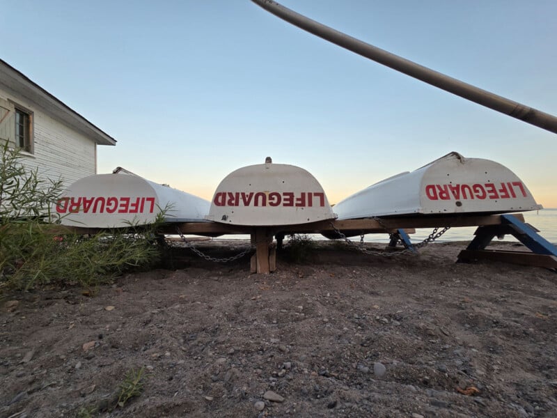 Three white lifeguard rowboats are stored upside down on wooden supports on a sandy beach, with “LIFEGUARD” written in red letters on each boat. A building is on the left, and the water is visible in the background.