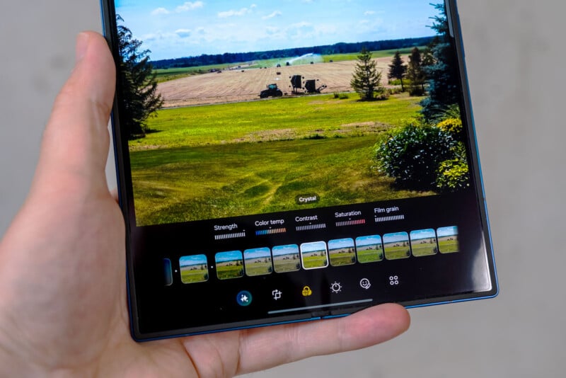 A hand holds a tablet displaying a landscape photo editing screen with image filters and adjustment options, showing a green field with trees and farmland in the background.