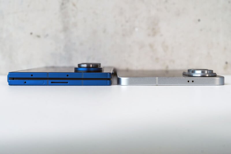 Two smartphones are shown side by side on a white surface, viewed from the edge. One phone is blue, the other silver, both with protruding rear cameras. The background is a plain, light-colored wall.