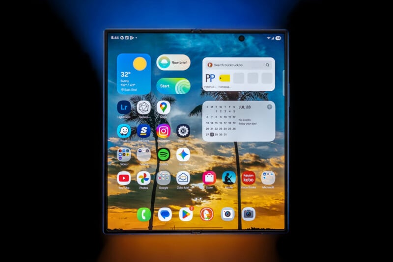 A foldable smartphone screen displays various app icons and widgets over a tropical beach sunset wallpaper with palm trees. The time is 5:46, and the date is July 28. The device is surrounded by a blue and orange gradient.