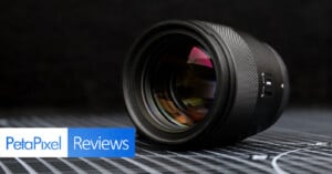 A camera lens is placed on its side on a grid-patterned surface, with "PetaPixel Reviews" text in the lower left corner against a dark, blurred background.