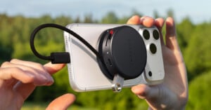 A person holds a smartphone outdoors with a circular Satechi device attached to the back, connected by a short cable, against a blurred background of green trees.