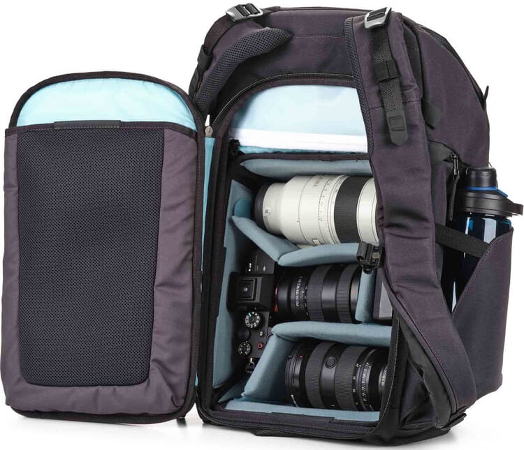 A black camera backpack is open, revealing padded compartments with a camera body and several camera lenses securely stored inside. A water bottle is held in an outer side pocket.