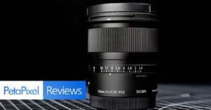 A Sigma 12mm f/1.4 camera lens is displayed upright on a grid-patterned surface with a dark background. The "PetaPixel Reviews" logo appears in the bottom left corner.