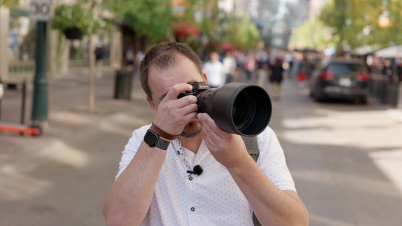 A person in a short-sleeved white shirt is taking a photo with a large camera on a city street, with people and trees blurred in the background.