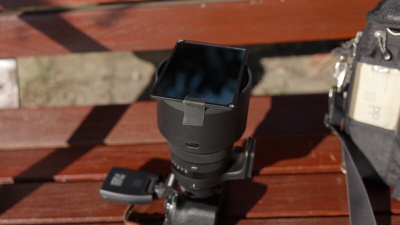 A camera lens with a square filter taped to the front is resting on a wooden bench. Part of a camera bag with a name tag is visible on the right side of the image.