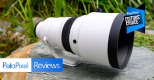 A large white telephoto camera lens is displayed outdoors on a rock, surrounded by grass. The image features "PetaPixel Reviews" and "PetaPixel Editors' Choice" badges.