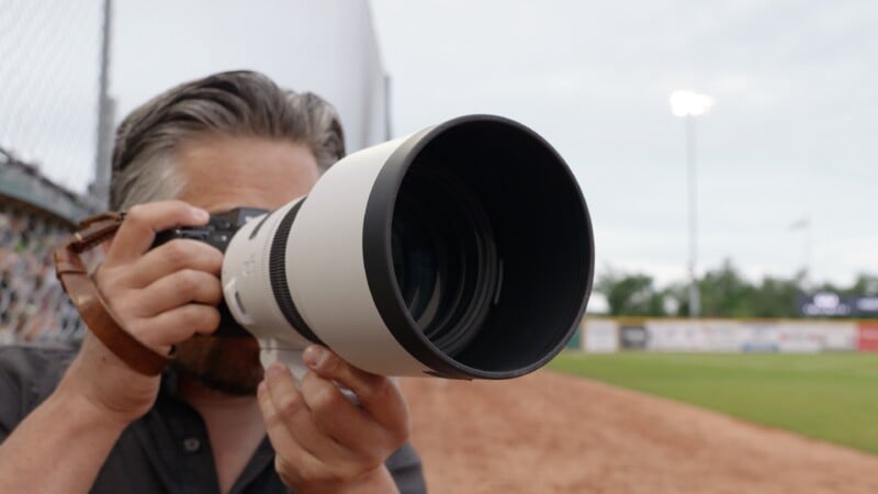 A person with dark hair is holding a camera with a large telephoto lens, aiming it toward a sports field, with a baseball diamond and outfield fence visible in the background.