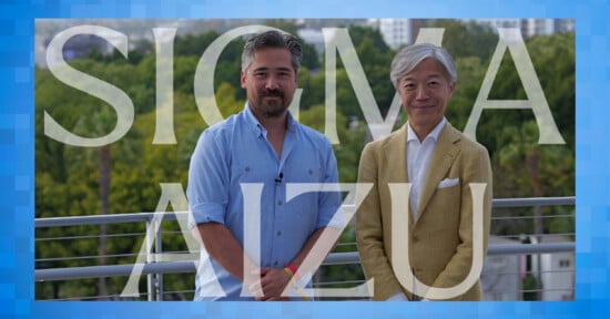 Two men stand side by side outdoors in front of greenery, one wearing a light blue shirt and the other a light brown suit. Large translucent text reads "SIGMA AIZU" across the image.