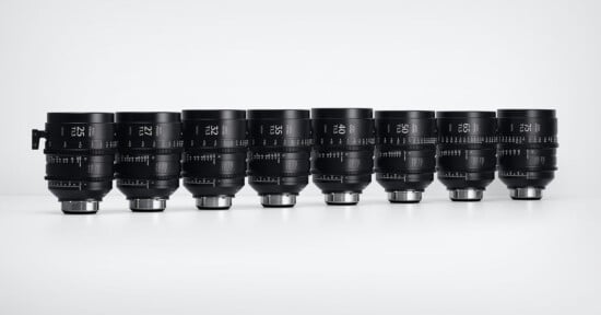 Seven black cinema camera lenses of varying sizes are lined up side by side on a white surface against a plain, light gray background. Each lens features white markings and numbers indicating specifications.