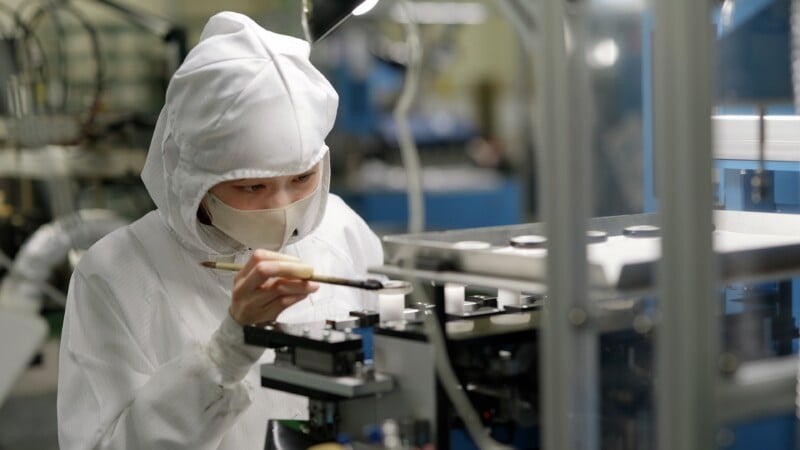 A person in a cleanroom suit and mask uses a brush to work on precision equipment in a laboratory or manufacturing facility, surrounded by machinery and technical instruments.