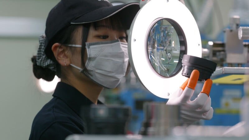 A person wearing a cap, face mask, and gloves inspects a camera lens under a bright circular light in a laboratory or factory setting.