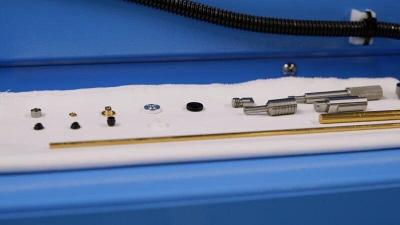 Small metal and plastic components, rods, and cylindrical parts are neatly arranged on a white cloth with a blue background, possibly as part of a mechanical or engineering assembly.