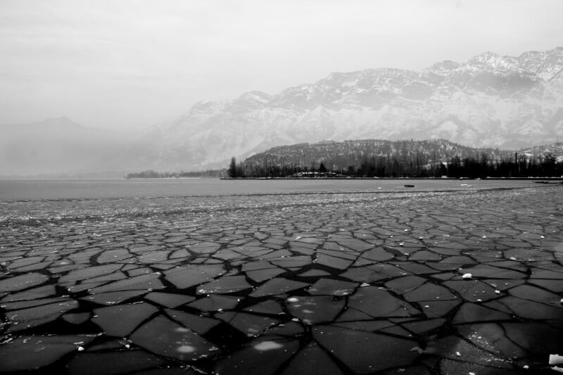A black and white photo of a frozen lake with cracked ice patterns in the foreground, leafless trees along the shore, and snow-covered mountains in the background under a cloudy sky.