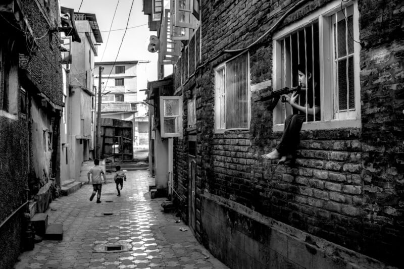 Black-and-white photo of a narrow alley where a child sits in a window, playing with a toy gun, while two adults and another child walk away along the wet, tiled street between tall, aged buildings.
