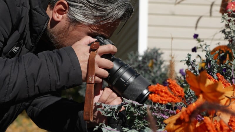 A person with gray hair and a beard, wearing a black jacket, is taking a close-up photo of orange and purple flowers with a professional camera outdoors on a sunny day.