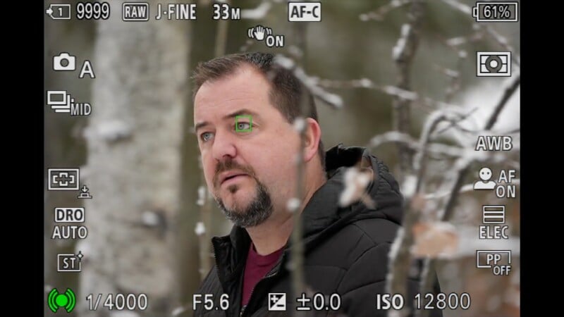 A man with short dark hair and a beard stands in a snowy forest, wearing a black jacket. Camera settings and focus indicators overlay the image, suggesting it is seen through a camera viewfinder.