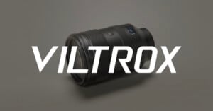 A black camera lens lies on a light gray surface with the word "VILTROX" in large, bold white letters displayed across the image.
