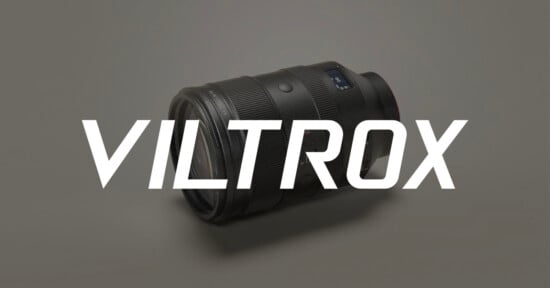 A black camera lens lies on a light gray surface with the word "VILTROX" in large, bold white letters displayed across the image.