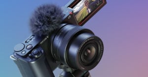 A Sony digital camera with a fluffy microphone attachment is mounted on a tripod. The flip-out screen displays a person taking a selfie outdoors with green fields in the background.