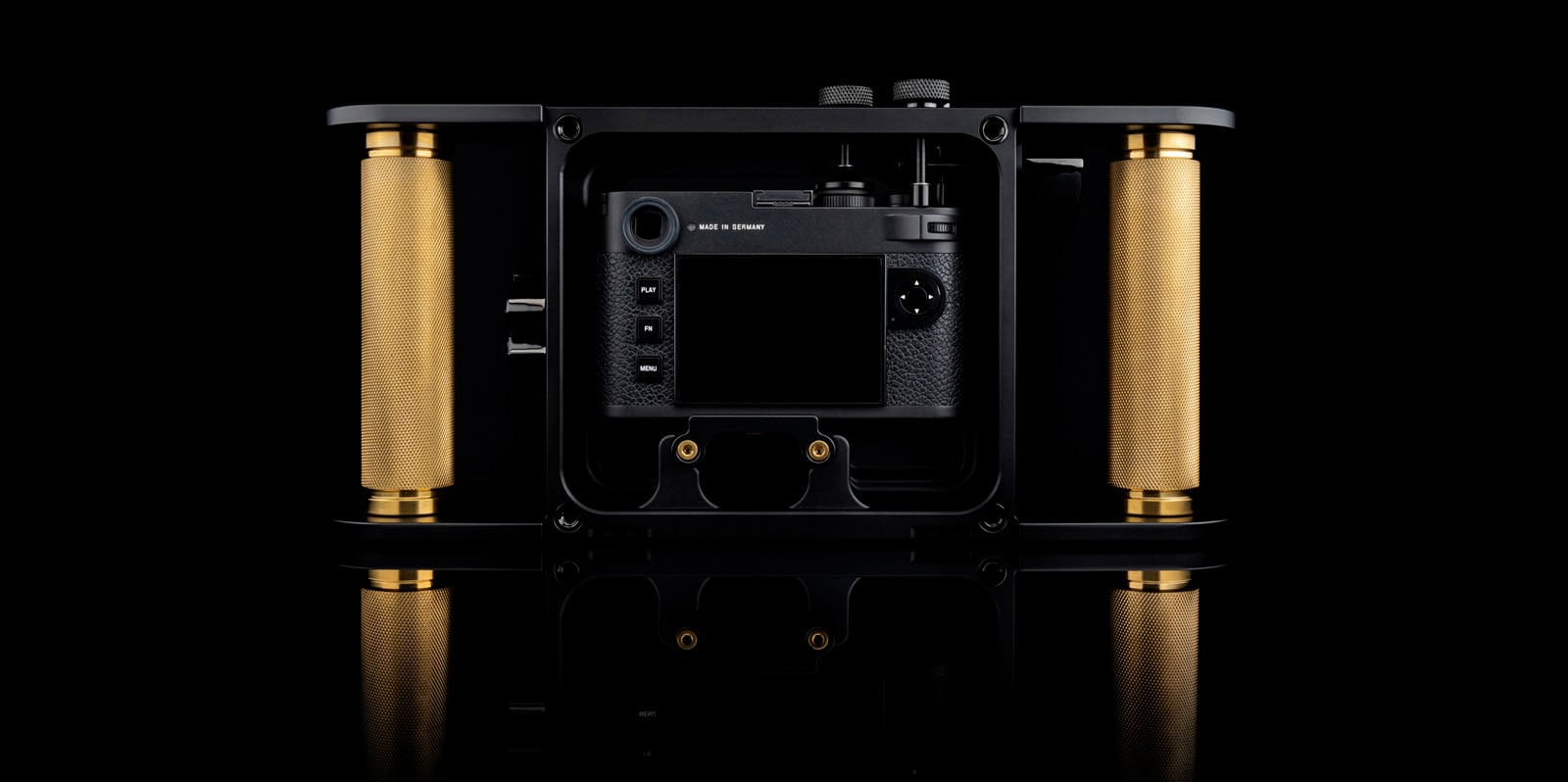 A professional camera rig with gold cylindrical grips on each side, featuring a mounted black camera centered within a rectangular black frame, displayed against a black background with a reflective surface.