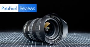 A wide-angle camera lens lies on a grid-patterned surface with a black background. The top left corner displays the "PetaPixel Reviews" logo in blue and white.