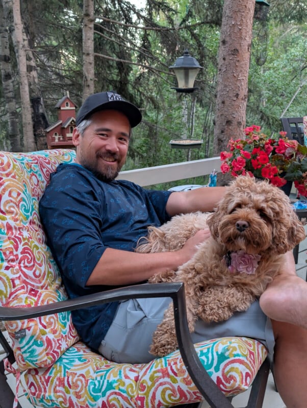 A man with a beard and baseball cap smiles while relaxing on a colorful patio chair, holding a curly-haired dog in his lap. Flowers and trees are visible in the serene outdoor background.