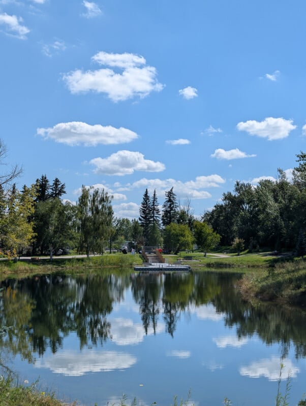 A calm pond reflects blue sky and fluffy clouds, surrounded by green trees and grass. A small wooden dock extends into the water, with a few scattered people in the distance under a sunny sky.