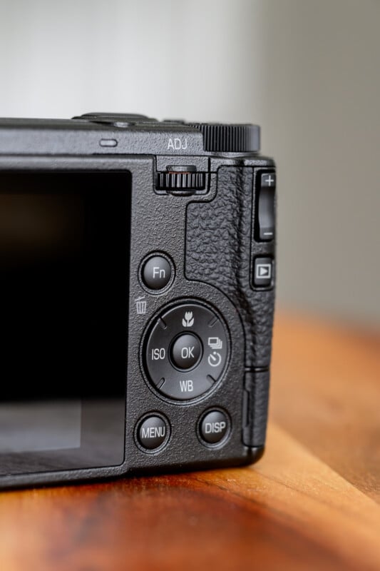 Close-up view of the back control panel of a digital camera, showing buttons such as ISO, WB, MENU, and DISP, along with a textured grip and adjustment dial on a wooden surface.