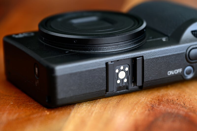 A close-up of a black digital camera on a wooden surface, focusing on its exposed hot shoe mount near the ON/OFF switch.