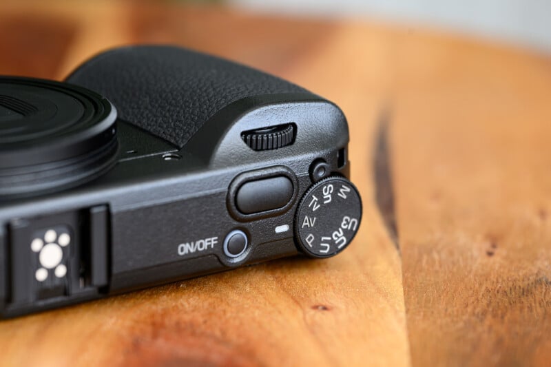 Close-up of a digital camera’s mode dial and power switch, resting on a wooden surface. The dial displays various shooting modes, including P, Tv, Av, M, and custom settings.