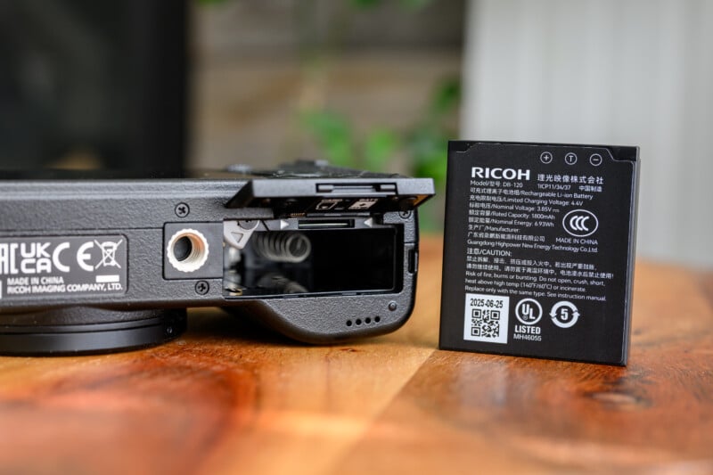 A Ricoh camera with its battery compartment open sits on a wooden surface next to a Ricoh rechargeable lithium-ion battery standing upright, displaying the battery's label and specifications.