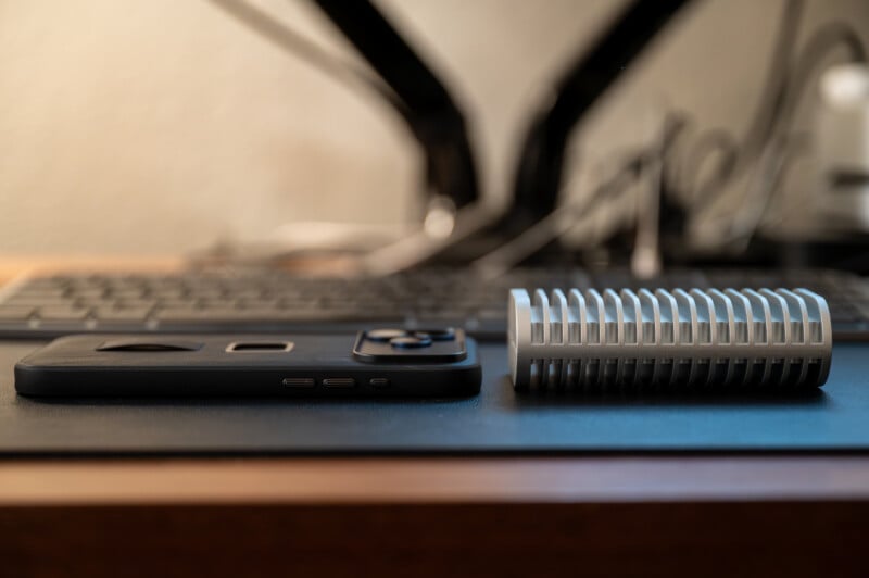 A black smartphone and a cylindrical, silver metal object with ridges lie side by side on a desk, with a blurry computer keyboard and monitor stand in the background.
