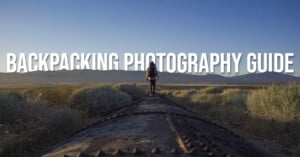A backpacker walks on top of a rusty pipeline surrounded by bushes with mountains and a clear blue sky in the background. Large white text reads "BACKPACKING PHOTOGRAPHY GUIDE.