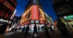 Crowds walk past brightly lit stores with large neon signs and advertisements in Japanese on a busy city street at night in an urban area, likely in Japan.