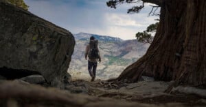 A hiker with a large backpack walks toward a scenic mountain vista, framed by a tree and a large rock, under a partly cloudy sky.