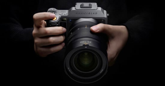 A person holding a Hasselblad camera with both hands against a dark background, with the camera lens and controls clearly visible.