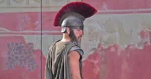 A man dressed as an ancient Roman soldier stands in profile, wearing a metal helmet with a large red plume and a cape, in front of a faded red wall with decorative patterns.