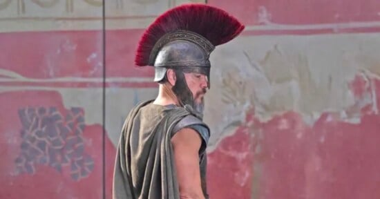 A man dressed as an ancient Roman soldier stands in profile, wearing a metal helmet with a large red plume and a cape, in front of a faded red wall with decorative patterns.