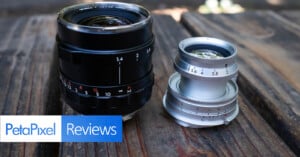 Two camera lenses sit on a wooden surface under natural light. The left lens is black with a red marking, and the right lens is silver. The image features a "PetaPixel Reviews" label in the bottom left corner.