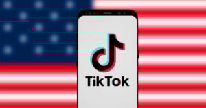 A smartphone displays the TikTok logo on its screen, positioned in front of a blurred American flag in the background.