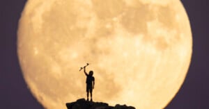 A person stands on a rocky hilltop holding an object above their head, silhouetted against a giant, bright full moon in the background.