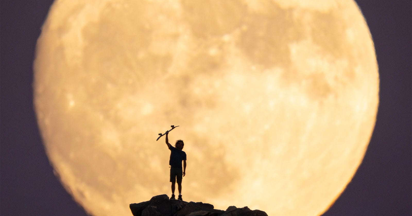 A person stands on a rocky hilltop holding an object above their head, silhouetted against a giant, bright full moon in the background.